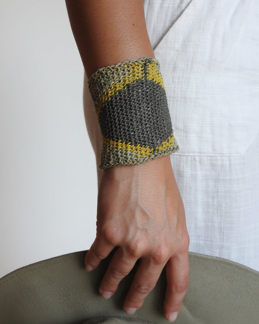 A close-up of a person's forearm and hand wearing a knitted wrist cuff in gray and yellow, resting against a white fabric-covered surface.