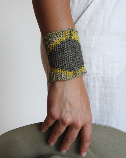 A close-up of a person's forearm and hand wearing a knitted wrist cuff in gray and yellow, resting against a white fabric-covered surface.